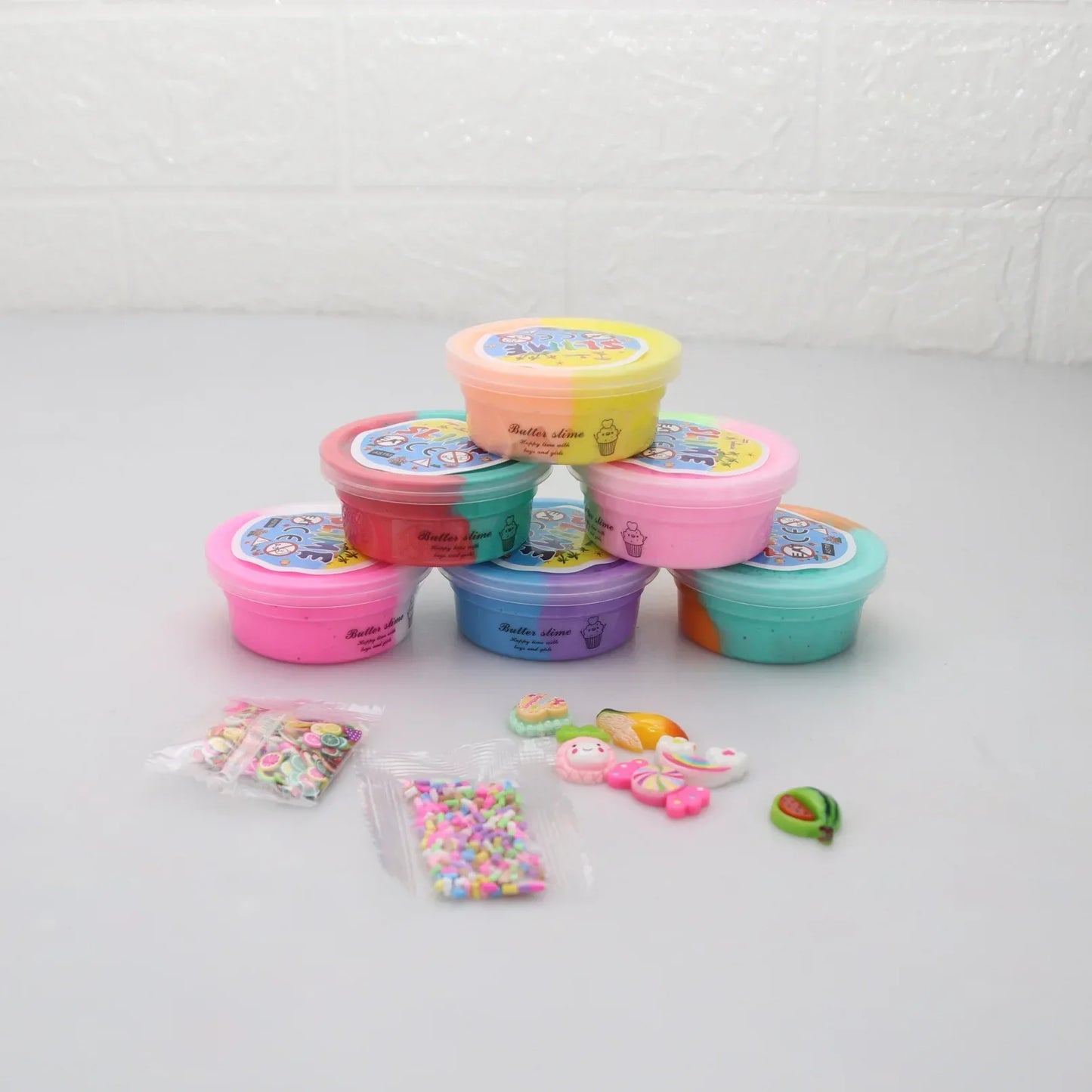 A stack of colorful slime containers, including pink, purple, and yellow, against a light background. The Slime Fluffy Kit for Enfant features miniature charms and sprinkles for decoration—a perfect DIY set that’s safe and non-toxic for all ages.