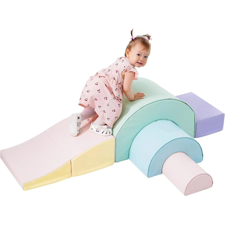 A toddler climbs on the Rainbow Soft Play Set—soft climbing toys for toddlers with foldable slide and soft-hued foam blocks—next to a classic wooden toy, set against a white background.