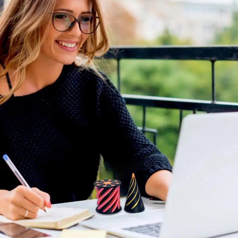 A smiling woman with glasses works on a laptop at an outdoor office desk, writing in a notebook. Two Impossible Cone Sensory Fidgets Toys are placed beside her as desktop stress relief decor.