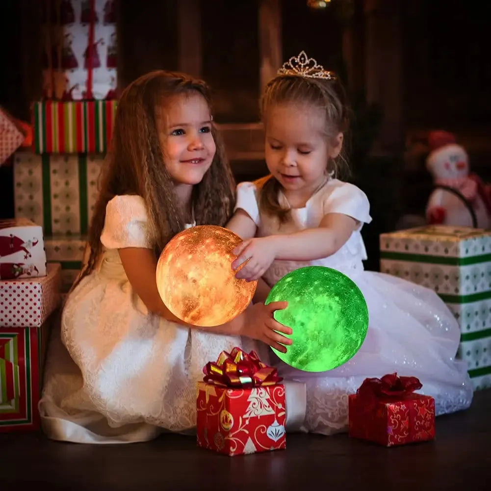 Two young girls in white dresses hold glowing lamps—a Celestial 3D Moon Lamp in orange and a Galaxy Lamp in green—amidst wrapped presents. They sit on a festive floor with a snowman decoration, adding magic to the scene.