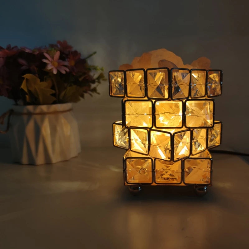 The Himalayan Salt Crystal LED Lamp, powered by USB, emits a warm glow that enhances the cozy ambiance of your room. It sits next to a white vase with pink and yellow flowers, inviting warmth and tranquility into the space.