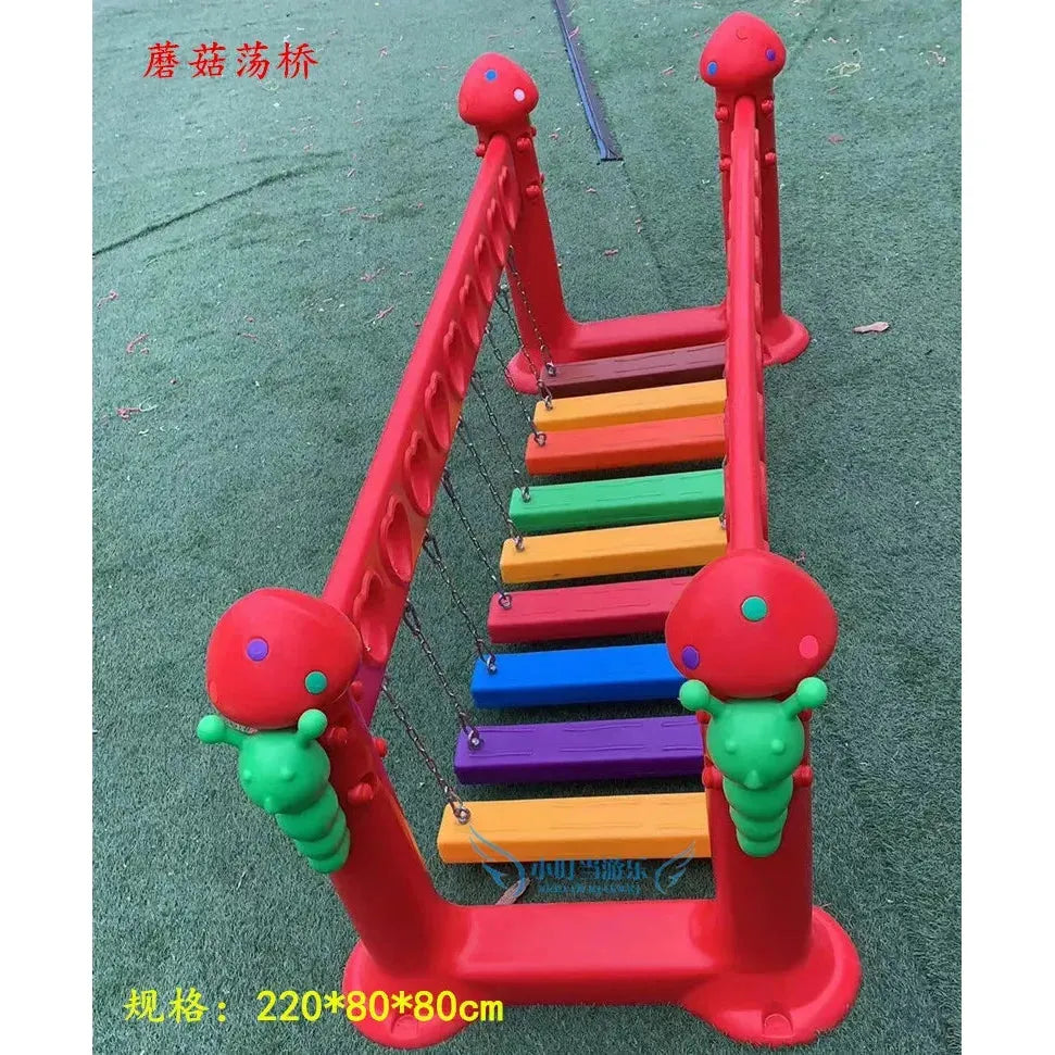 The outdoor toys feature a dark khaki childrens swinging bridge with decorative ladybug elements on red railings. The walkway is made up of multicolored steps set on green artificial turf.