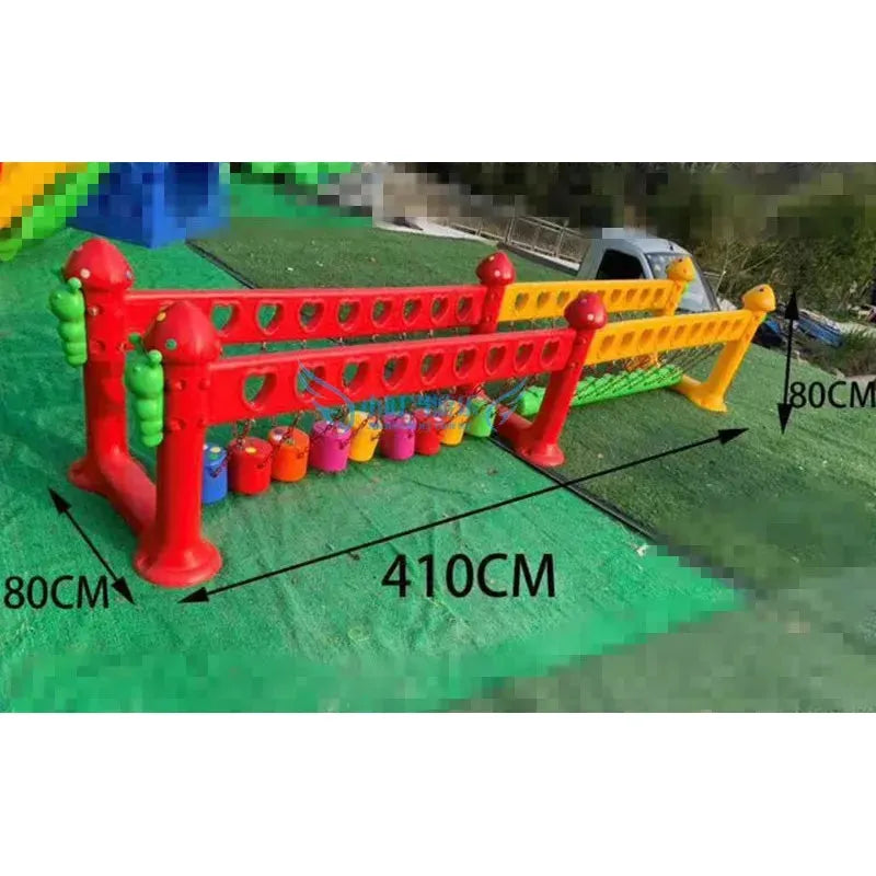 A green outdoor scene with chocolate-colored plastic barriers featuring heart-shaped cutouts. Two toy-filled containers hang from a red and yellow barrier. Dimensions: 410 cm length x 80 cm height, with a blurred background showcasing childrens swings, climbing nets, and swinging bridges.