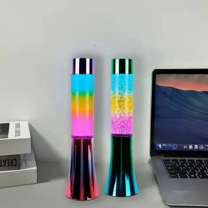Two 13-inch Color-Changing LED Glitter Lava Lamps sit on a desk by a laptop with an ocean screensaver. The vibrant pink, blue, yellow, and green hues of these decorative pieces enhance the charm of the setup, complemented by two neatly stacked books on the left.