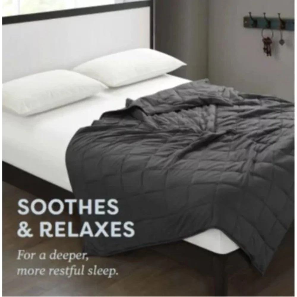 A neatly made bed showcases the Plush 20lb Soft Weighted Blanket, with a white frame, two plush pillows, and a premium dark gray design. The text reads, SOOTHES & RELAXES for deeper sleep. In the background, an Origin US-sourced shelf holds keys on hooks.