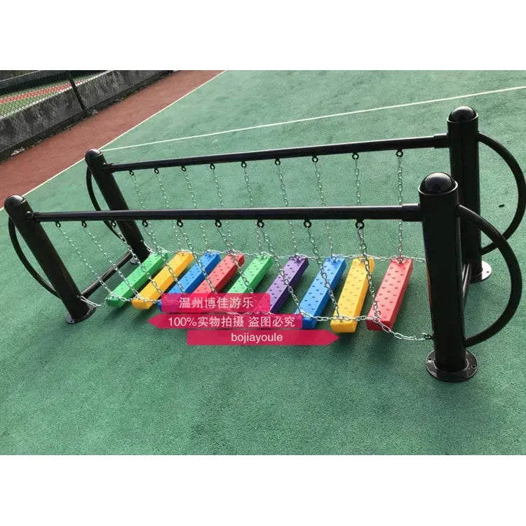 A colorful outdoor musical xylophone installation on a green surface, perfect for children aged 3-8. It features eight rainbow bars supported by black metal frames and chains, resembling a large metal toy. A tennis court fence is visible in the background.