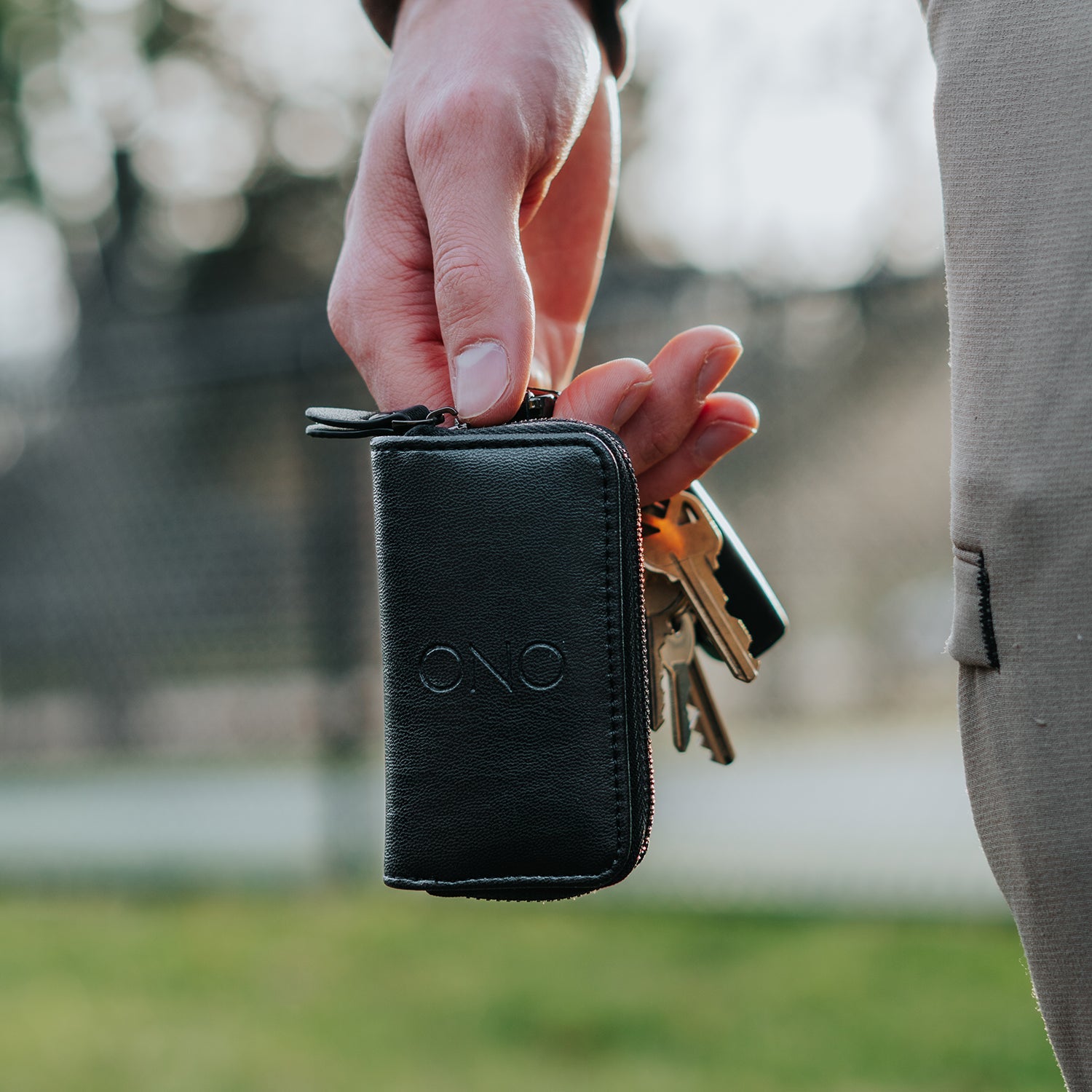 A person holds the ONO Roller Premium Keychain Case in black with several keys outdoors, against a blurred fence and greenery backdrop.
