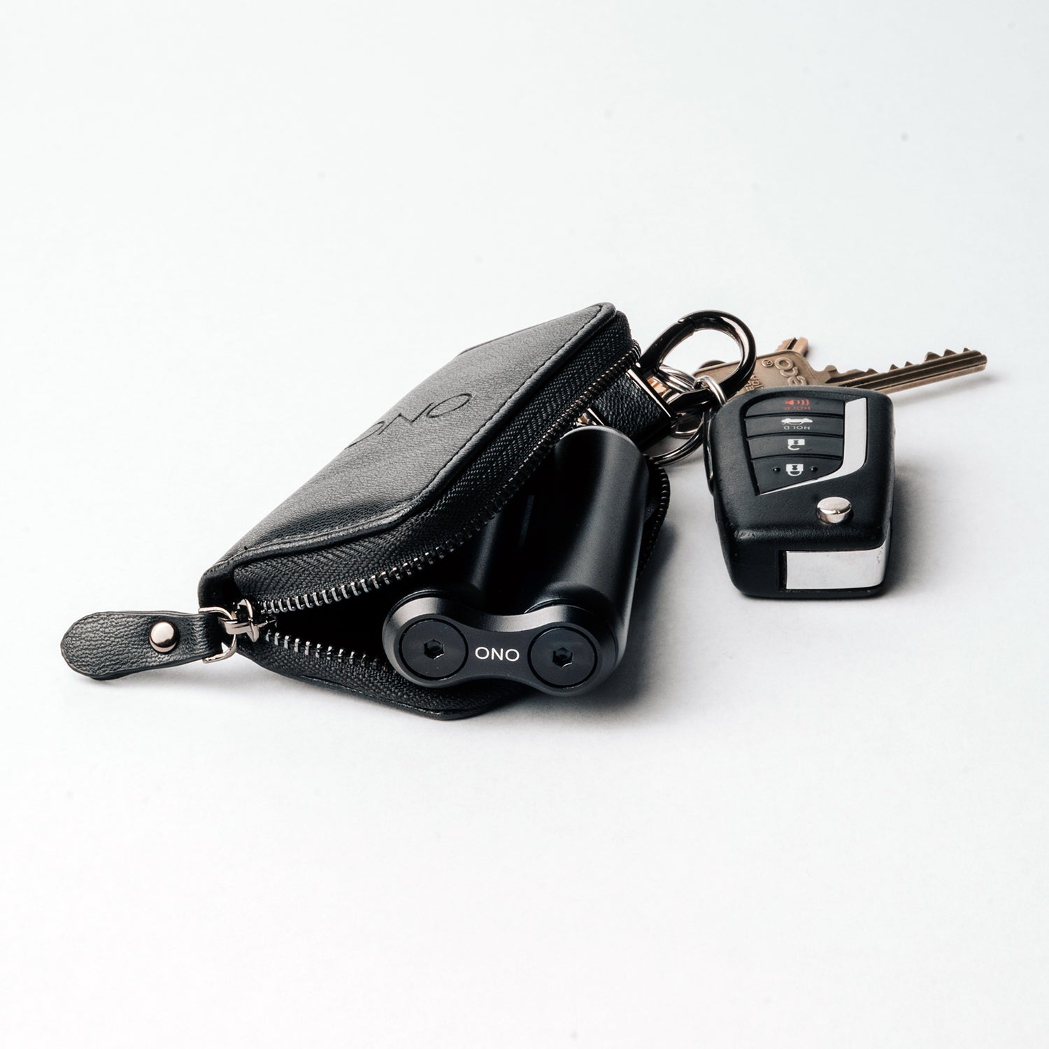The ONO Roller Premium Keychain Case in black, featuring a zipper and partially visible ONO wireless earbuds, is attached to a keyring with car and gold house keys against a white background.