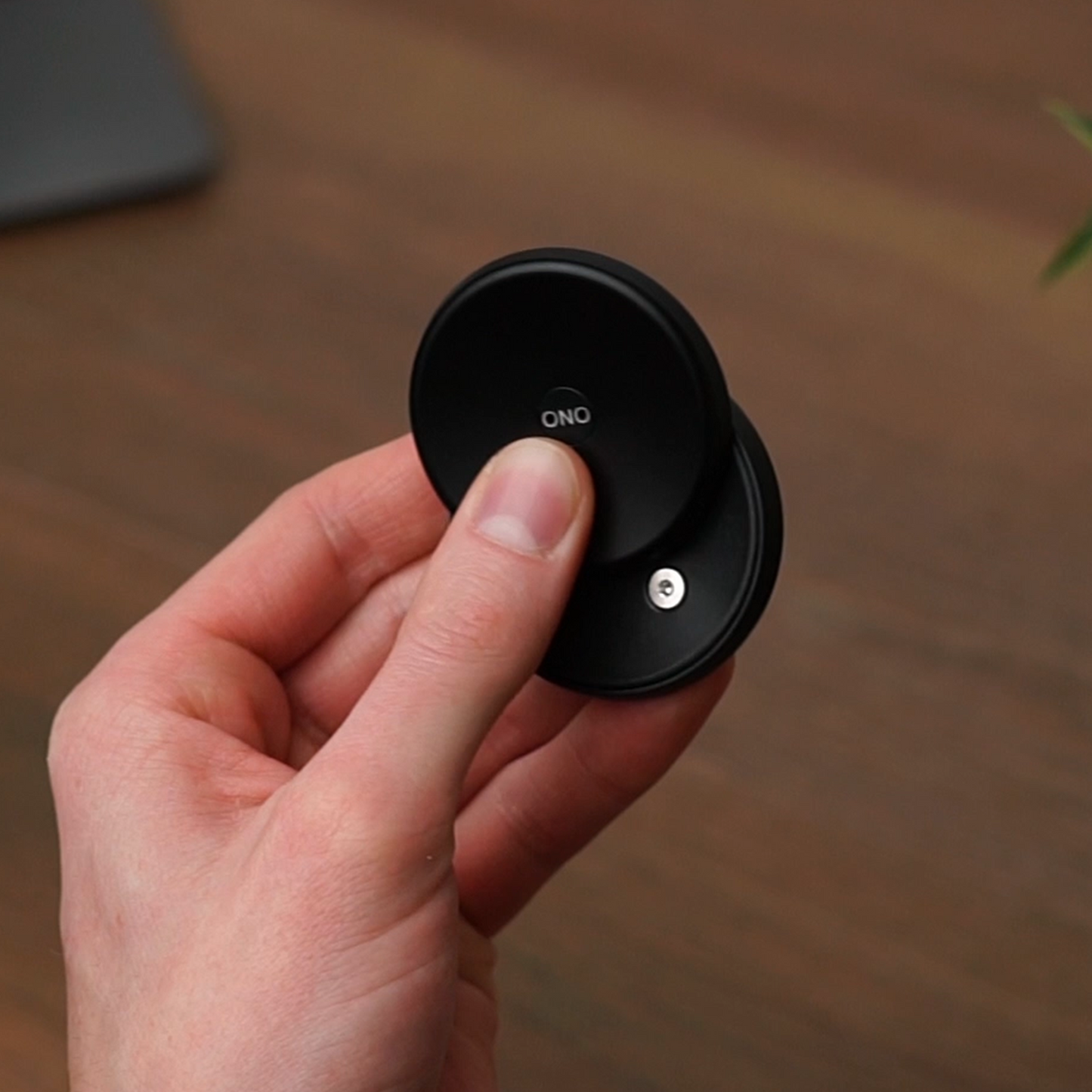 A hand holds a black ONO Slider Aluminum, a calming fidget toy, against a blurred wooden surface background.