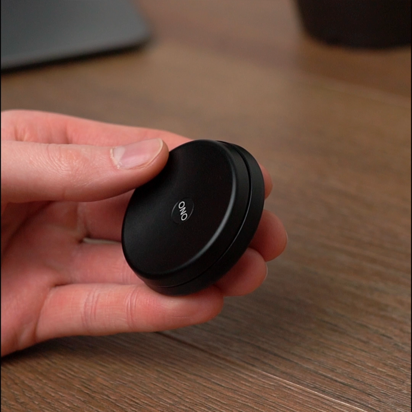 A hand holds a small, round, black ONO Slider Aluminum fidget toy against a wooden surface, with blurred objects in the background.