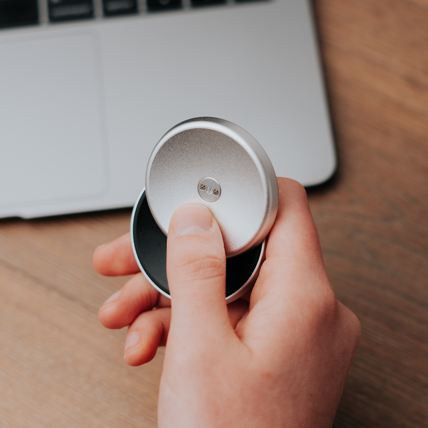 A hand holds the ONO Slider Aluminum, a calming fidget toy, above a wooden surface with part of a laptop keyboard visible in the background.