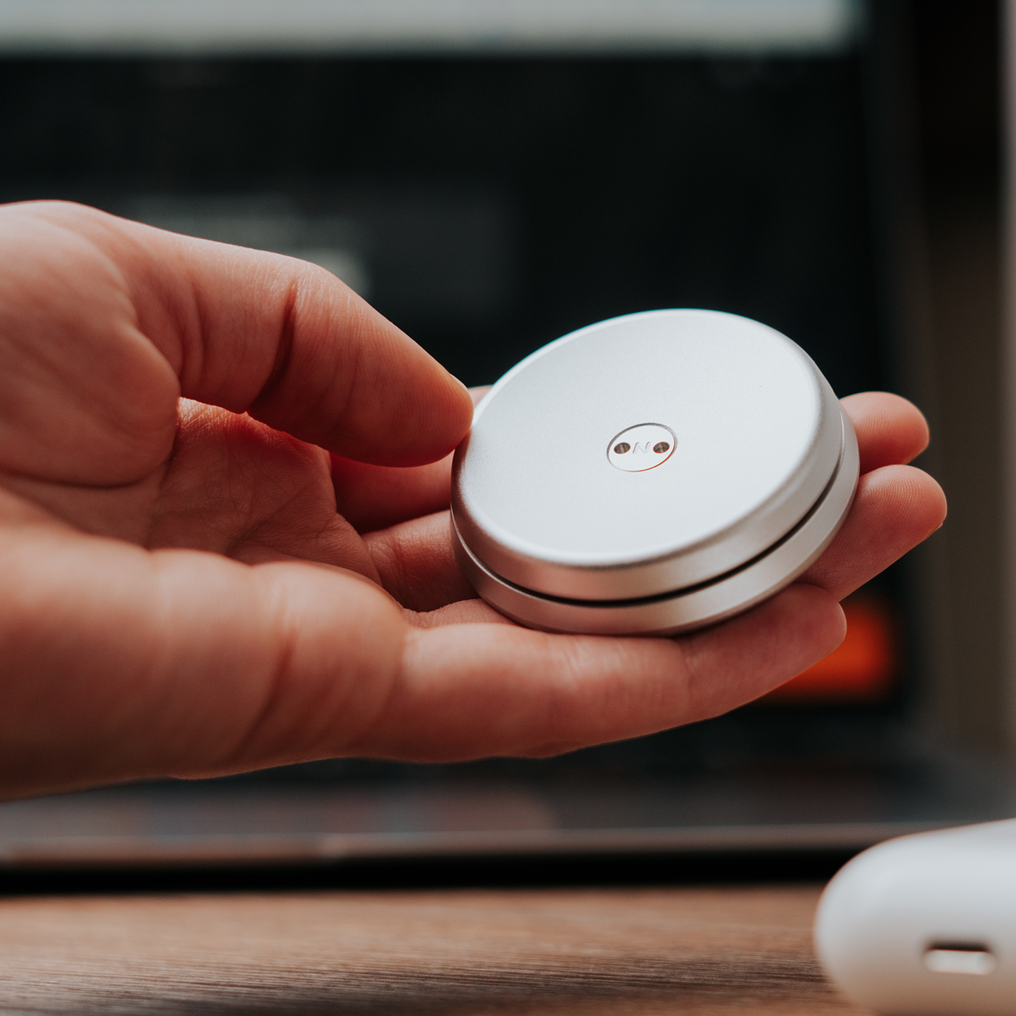 A hand holds a small, round ONO Slider Aluminum device with two metal contacts on top, set against a blurred background featuring a laptop and wooden surface.