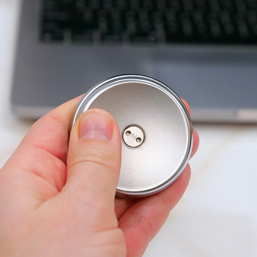 A hand holds a small, round ONO Slider Steel fidget toy with ONO engraved in the center, in front of a blurred laptop keyboard.