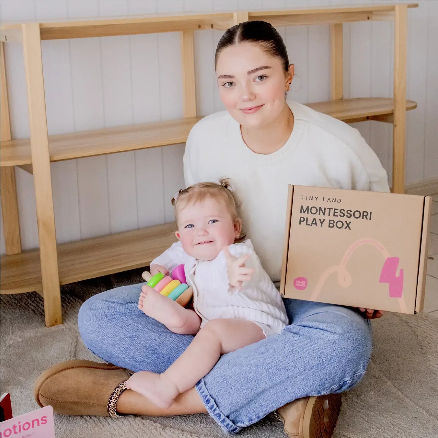 A woman sits on the floor with a smiling baby exploring Tiny Land® Montessori Toys for Infants (10–12 Months) from a cardboard box, in a bright, cozy room with wooden shelves in the background.