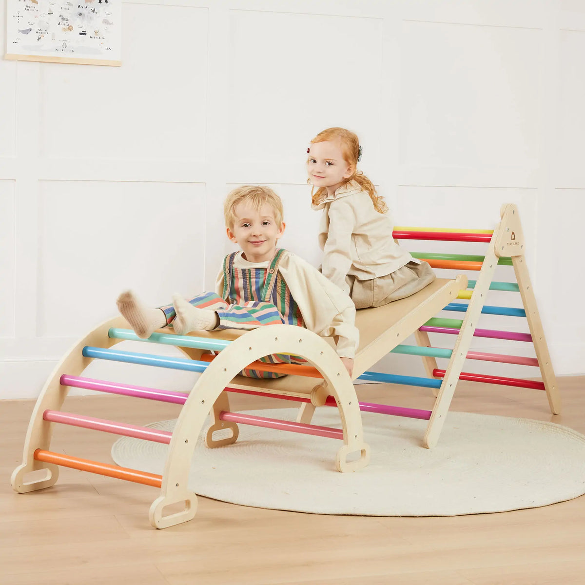 Two children sit on a wooden ramp connected to a 7-in-1 Montessori Climbing Set with Play Pad – Rainbow, featuring an arched climber and A-frame ladder with colorful rungs.