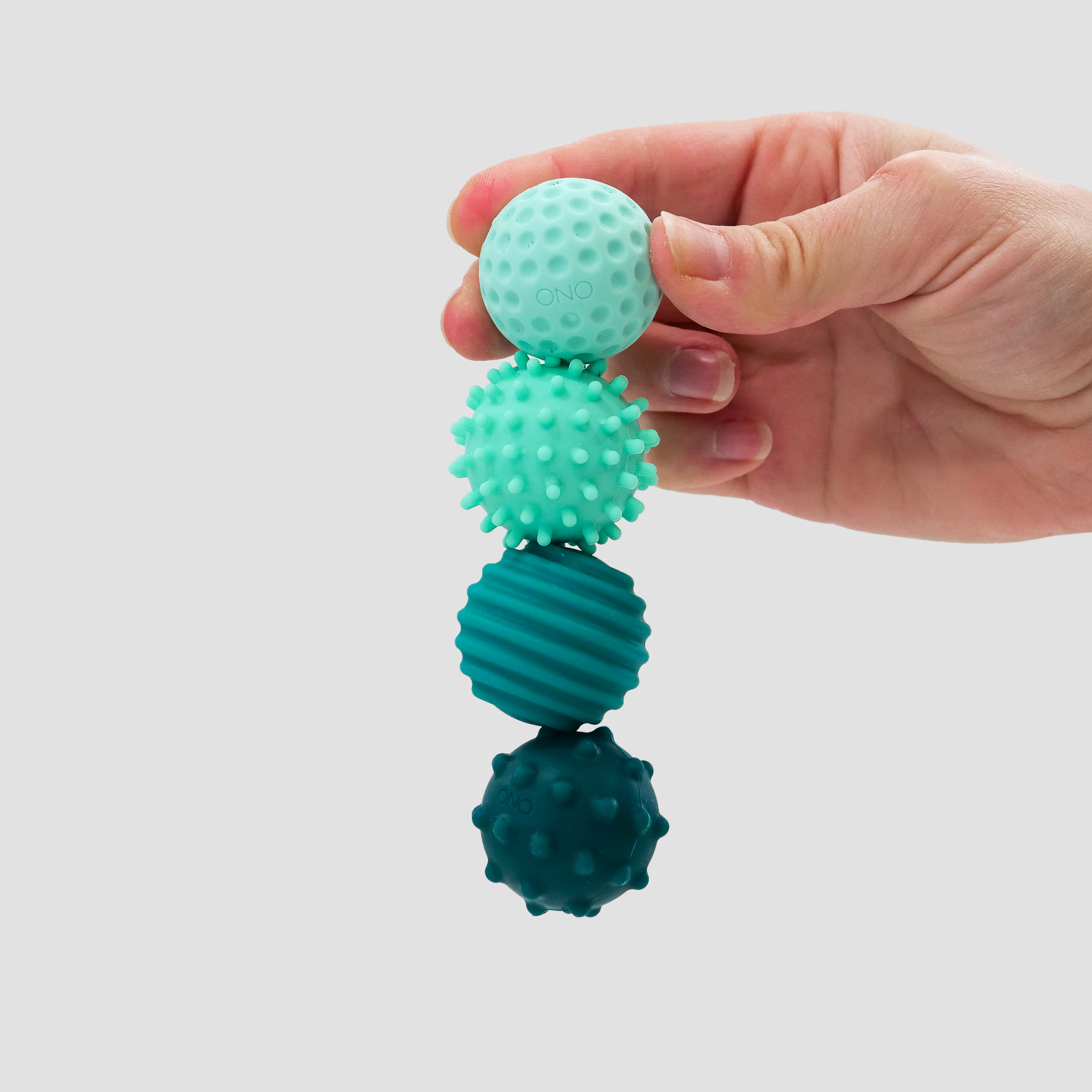 A hand holds four connected Signets—silicone-coated, magnetic balls in different teal shades and textures—against a light gray background, ideal for satisfying fidgeting and enhancing focus.