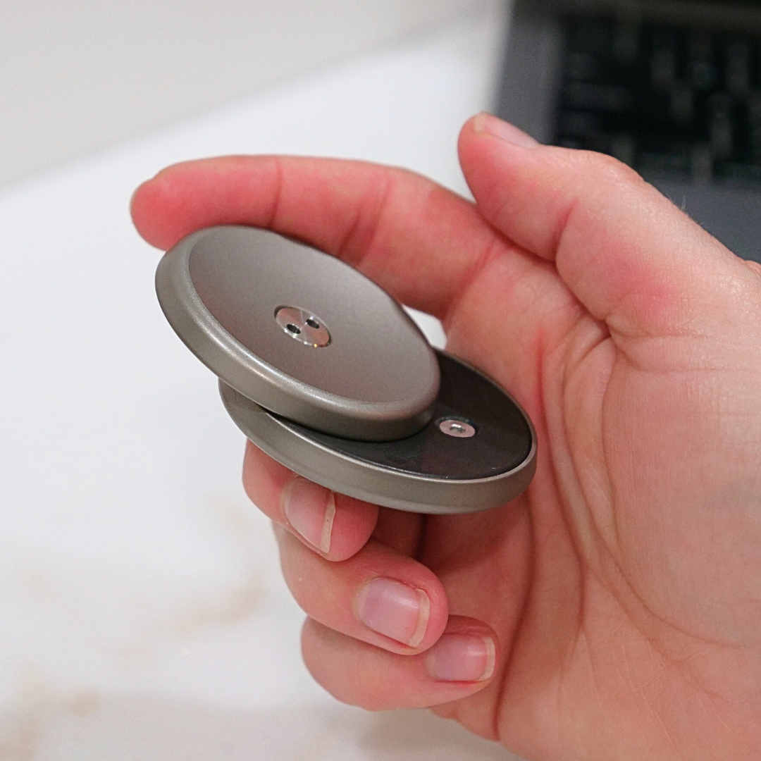A hand holds a round, metallic ONO Slider Titanium fidget toy with two central discs. In the background, part of a laptop rests on a desk.