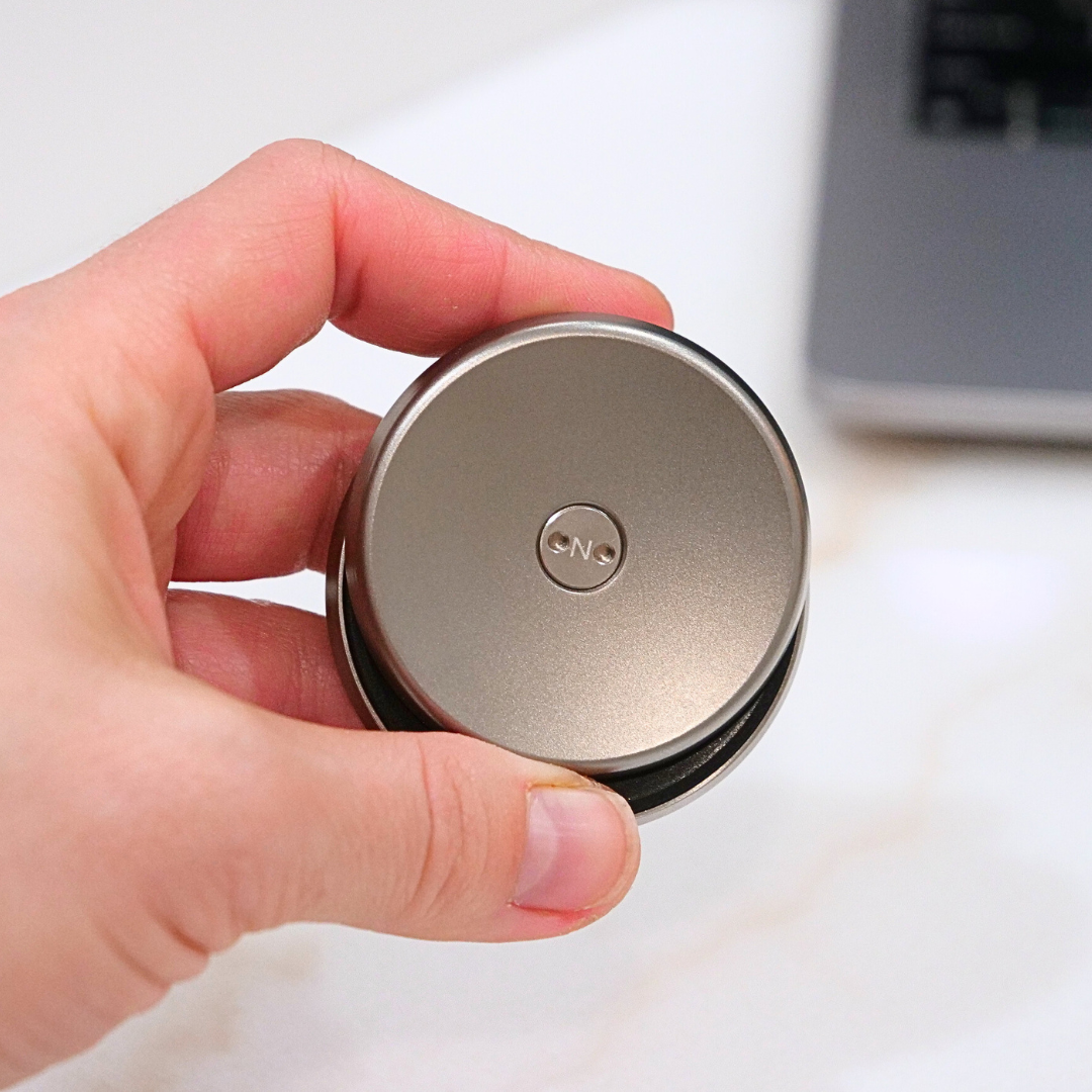 A hand holds the ONO Slider Titanium, a small, round fidget toy with ONO engraved on top; a laptop appears blurred in the background.