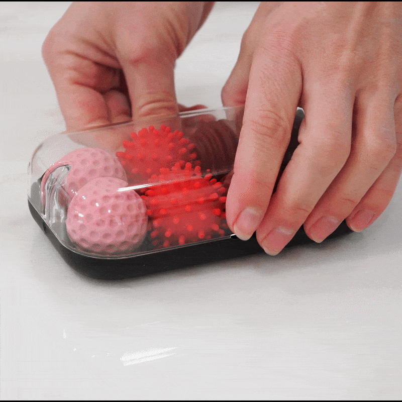 Two hands place a clear cover over a black tray holding Signets—two pink dimpled balls and two red spiky massage balls—providing a unique fidgeting experience that aids focus, all set on a white surface.