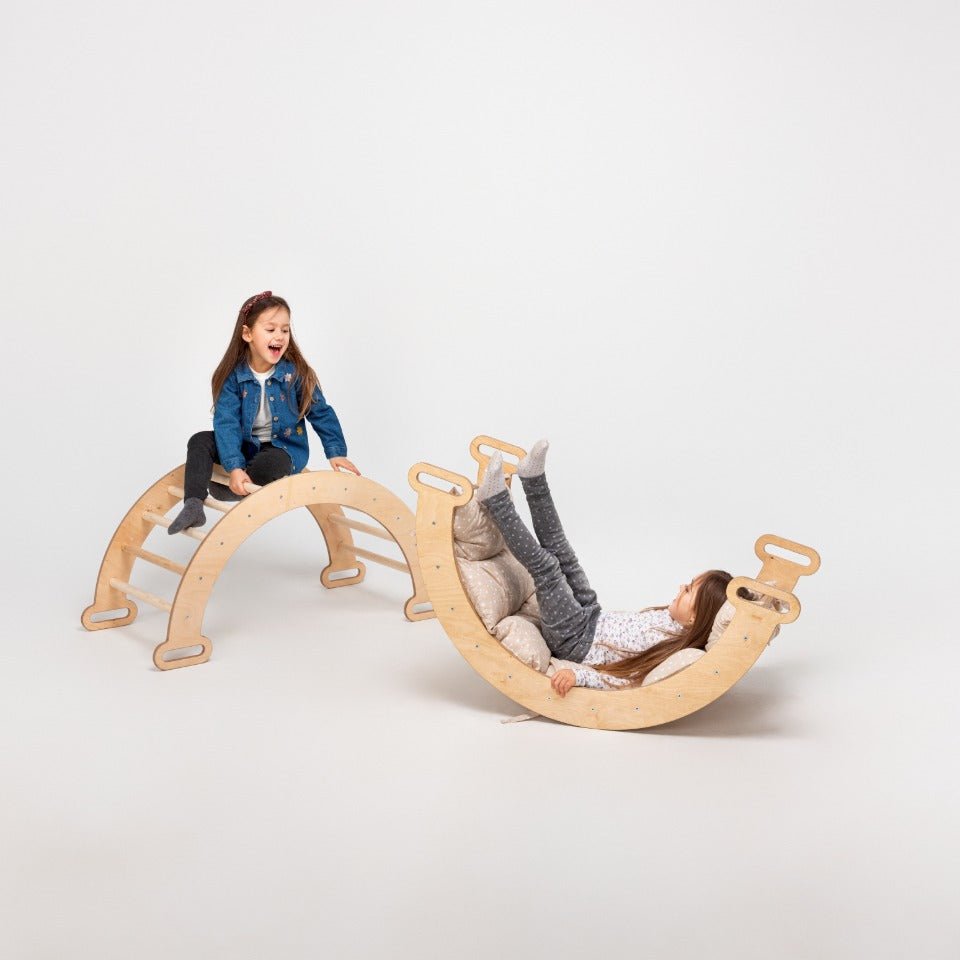 Two young girls engage in imaginative play; one smiles atop the beige Climbing Arch & Rocker Balance - Montessori Climbers for Kids 1-7 y.o., while the other reclines with her legs up on a curved balance board. The background is plain and light-colored.