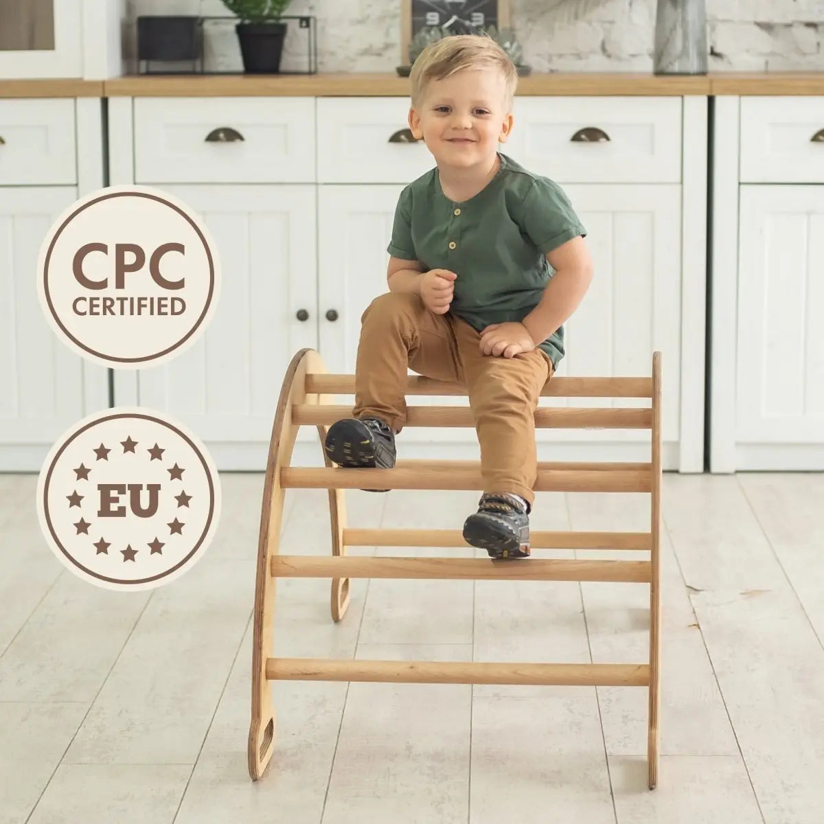 A young boy plays on a Climbing Arch & Rocker Balance - Montessori Climbers for Kids 1-7 y.o. – Beige in a bright kitchen with white cabinets and light wood countertop. Two circular badges nearby read CPC Certified and EU surrounded by stars.