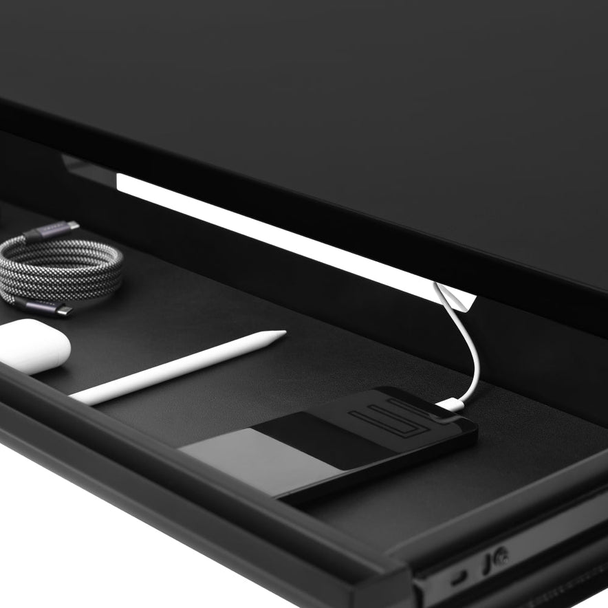 The Under Desk Drawer includes a coiled USB cable, a white stylus, a smartphone charging via USB, and a small white object, all organized for efficient under-desk storage.