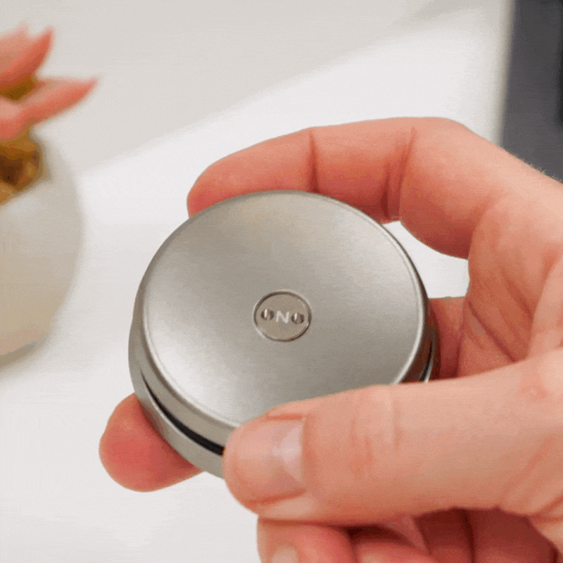 A hand spins a small, round ONO Slider Titanium fidget toy, engraved with “ONO” on top. A blurred plant appears in the background.