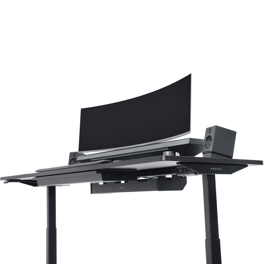 The Hexcal Elevate Standing Desk in black features height adjustability, a large curved monitor, two speakers, and a keyboard tray, all set against a plain white background.