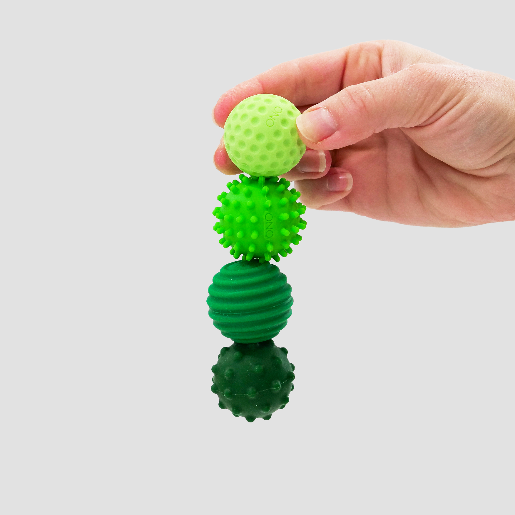 A hand holds three Signets—green, textured balls (dimpled, spiky, ridged)—stacked vertically against a light gray background, offering a unique fidgeting experience to aid focus.