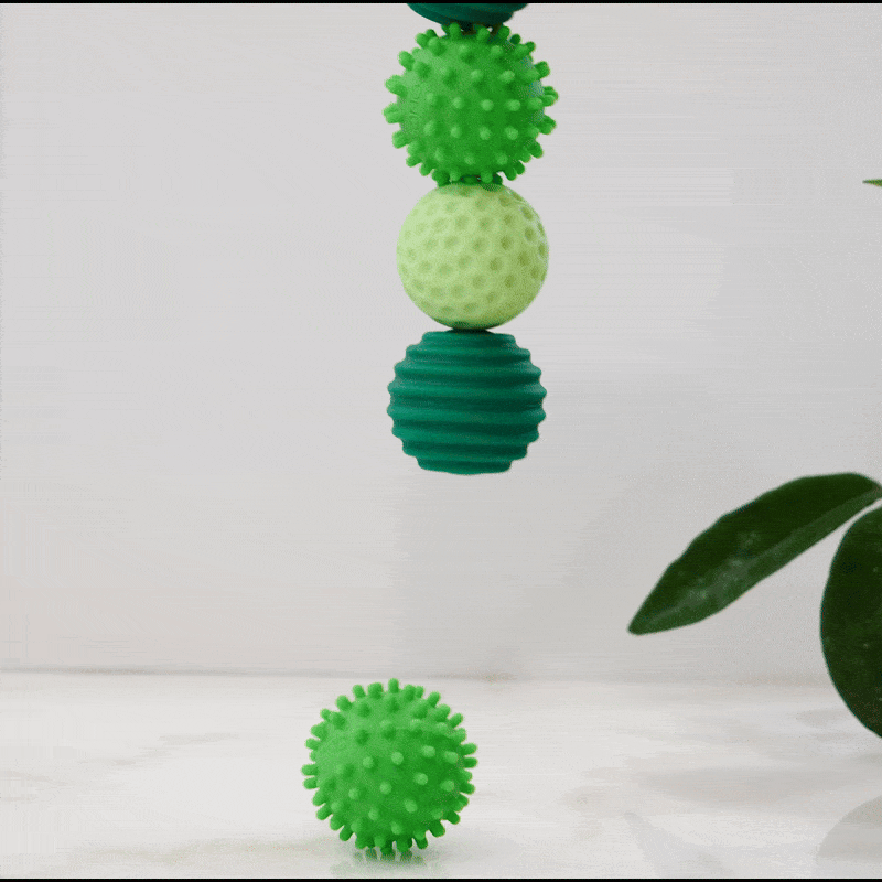 Four green Signets balls, stacked vertically, are shown; the bottom silicone-coated magnetic ball drops to the surface while three remain suspended. A green plant leaf is visible on the right against a light background.
