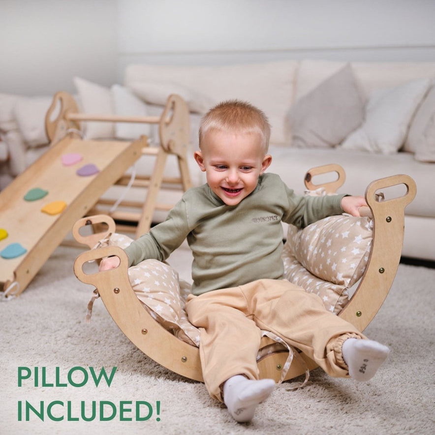 A smiling toddler sits in the 4in1 Montessori Climbing Set: Pikler Triangle, Arch, Slide Board, and Cushion for Toddlers (1–3 Years) in a cozy living room. Colorful grips are visible. Text reads PILLOW INCLUDED!.