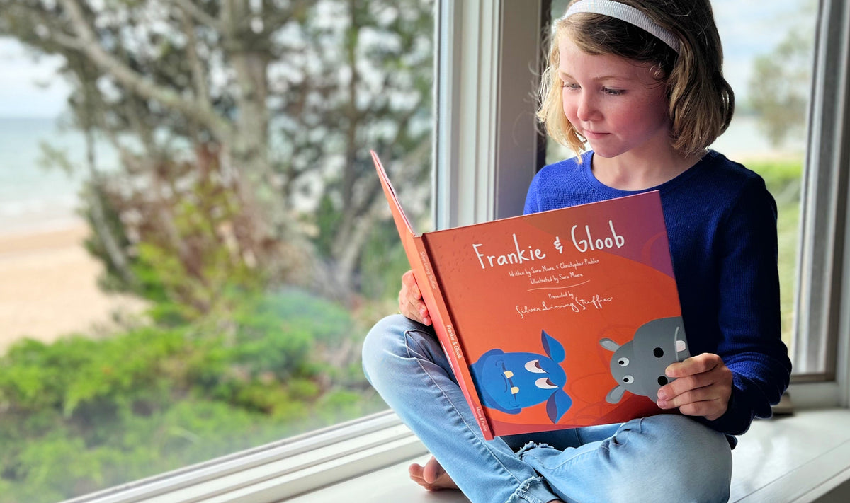 A young girl sits by a window reading the Frankie & Gloob – Picture Book, which features a blue character and a gray character on the cover.