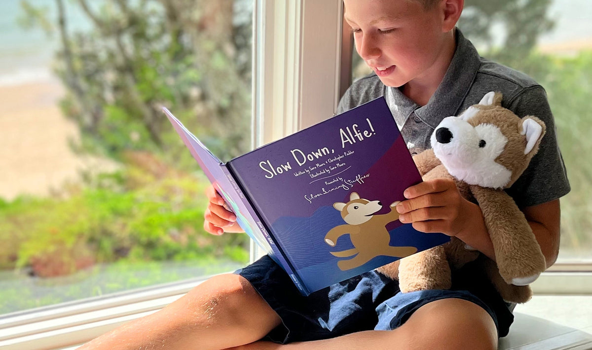 A boy sits by a window reading a book while holding his Alfie the Wolf Plush + Book.