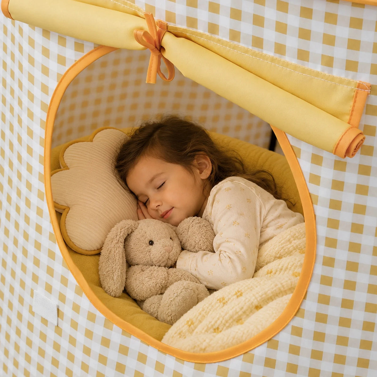 A young child sleeps inside a yellow and white checkered Starry Indoor Play Tent for Toddlers, snuggling a plush bunny toy next to a cloud-shaped pillow.