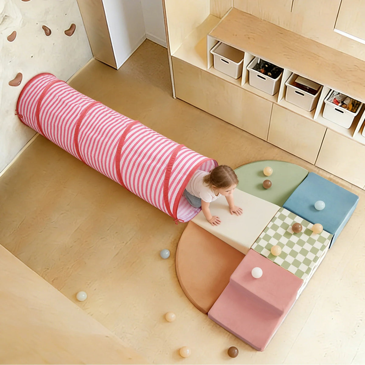 A toddler crawls out of a pink and white striped Adventure Crawl Tunnel (68″) onto a modular foam climbing set.