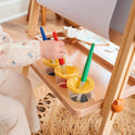A child dips a brush into red paint on the Tiny Land® Magnitales Easel for Kids tray, with cups of blue, yellow, and green paint and a roll of drawing paper—perfect for creative time on this height-adjustable easel.