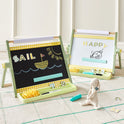 Two green 3-in-1 Adjustable Tabletop Art Easels stand on a patterned rug, one showing a chalkboard with a sailboat drawing and the other a whiteboard with the word HAPPY and a monkey sketch, including rulers, chalk, and a stuffed rabbit nearby.