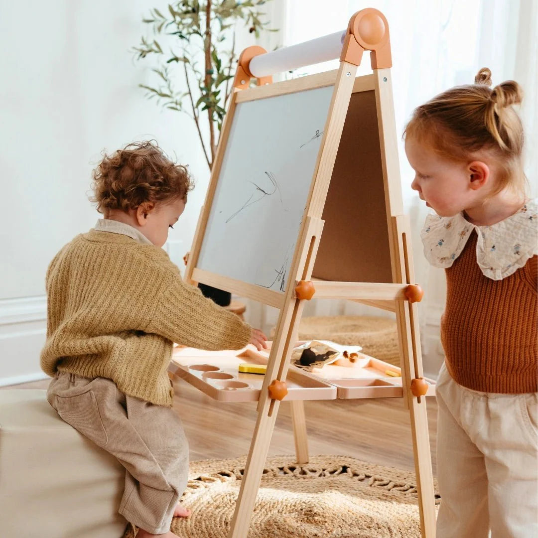 Two young children use the Tiny Land® Magnitales Easel for Kids in a bright, cozy room—one sits and draws while the other stands and watches. Art supplies are organized neatly on the easel’s tray.