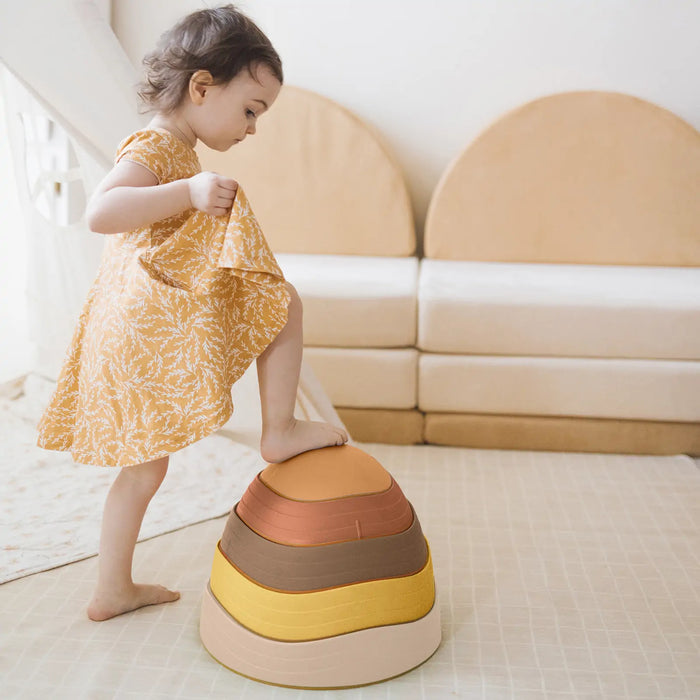 A toddler steps onto a Stacking Balance Stepping Stones – Sunset set of five nesting stones in cream, yellow, orange, and brown, forming a fun balance course.