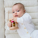A baby in a white, buttoned onesie lies on a padded, cream-colored surface, playing with a wooden rattle from the Tiny Land® Montessori Toys for Newborns (0–3 Months) sensory set.