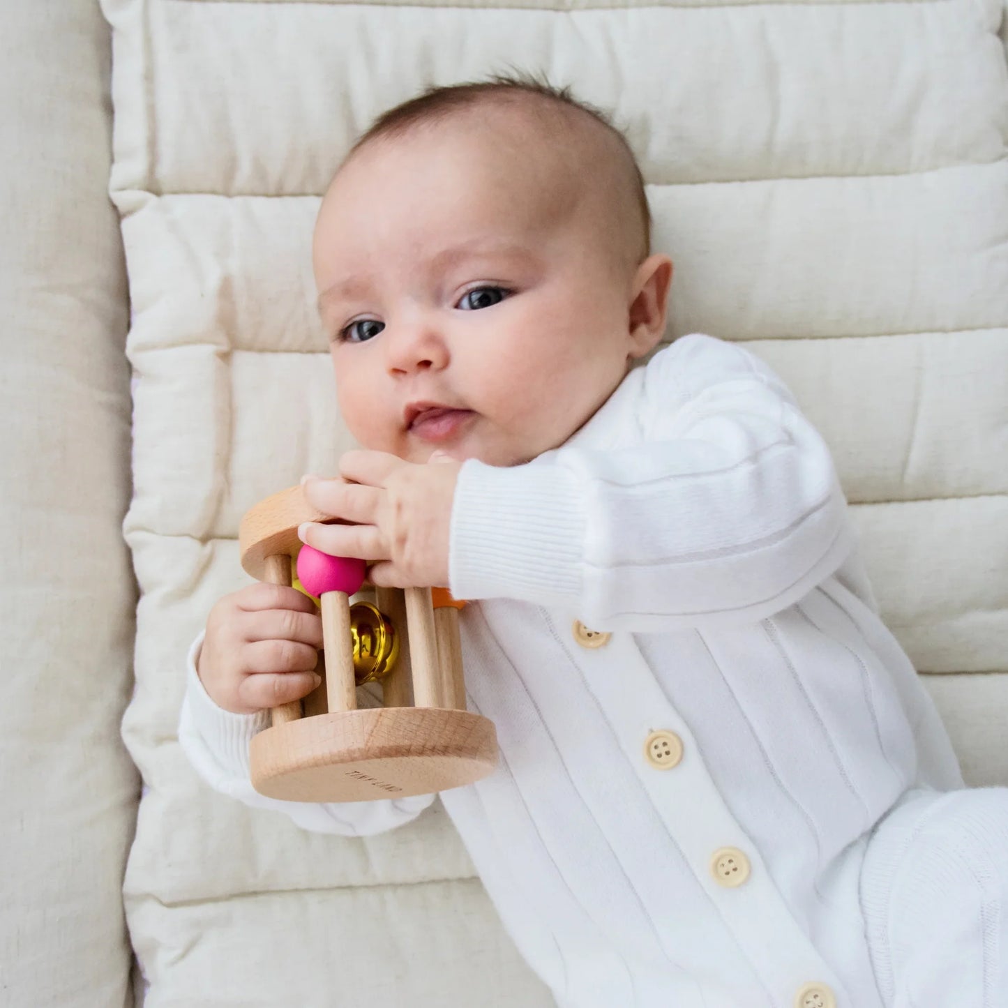 A baby in a white, buttoned onesie lies on a padded, cream-colored surface, playing with a wooden rattle from the Tiny Land® Montessori Toys for Newborns (0–3 Months) sensory set.