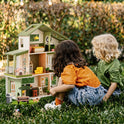 Two children sit on the grass playing with a Sweetwood Cottage Dollhouse (48-pc Furniture), a 1:12 scale wooden dollhouse furnished with miniature items.