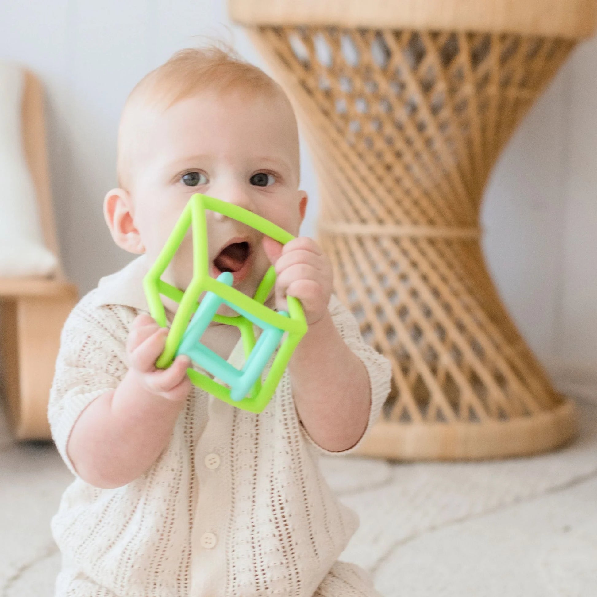 A baby in a cream knit outfit sits on a rug, exploring a green geometric toy from the Tiny Land® Montessori Toys for Newborns (4–6 Months) set—ideal for developing fine motor skills. Cozy furnishings and a wicker table are visible behind them.