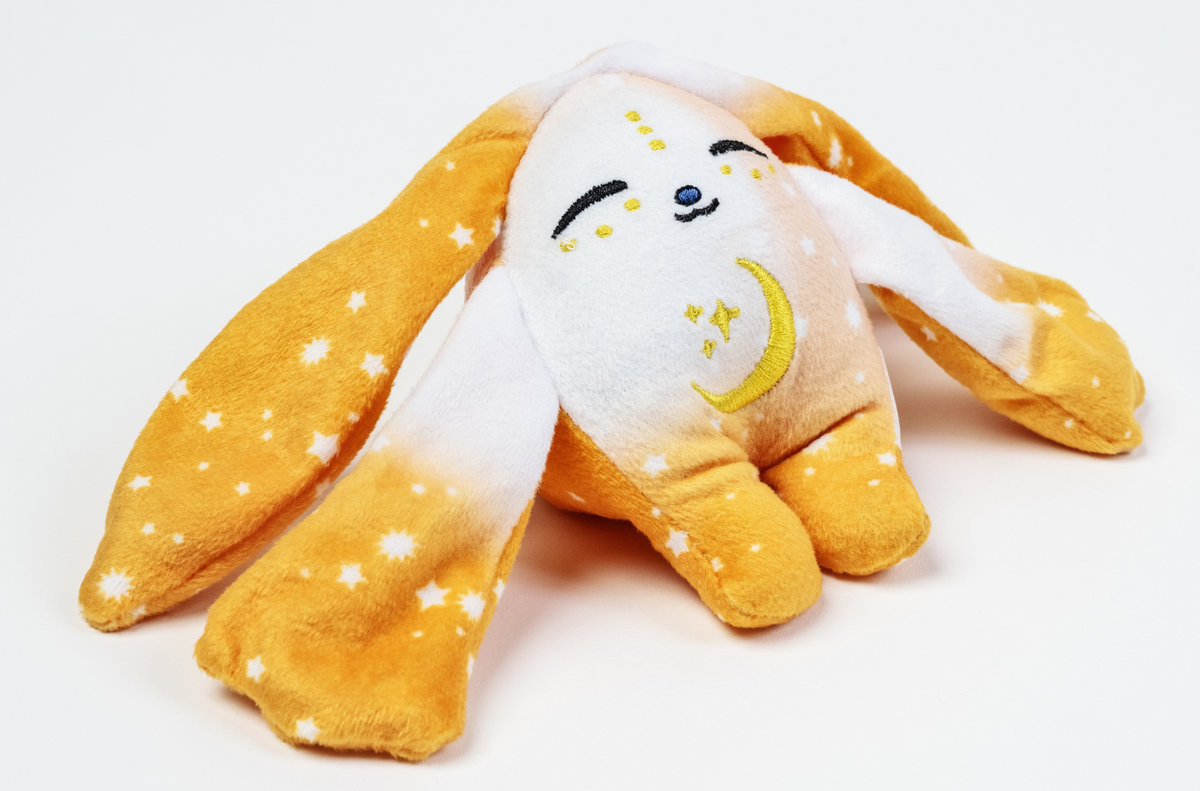 An Ori Mini Wrist Hugger – Magnetic Sensory Plush shaped like a rabbit, with long orange ears patterned with white stars and a white face featuring an embroidered yellow moon and stars.