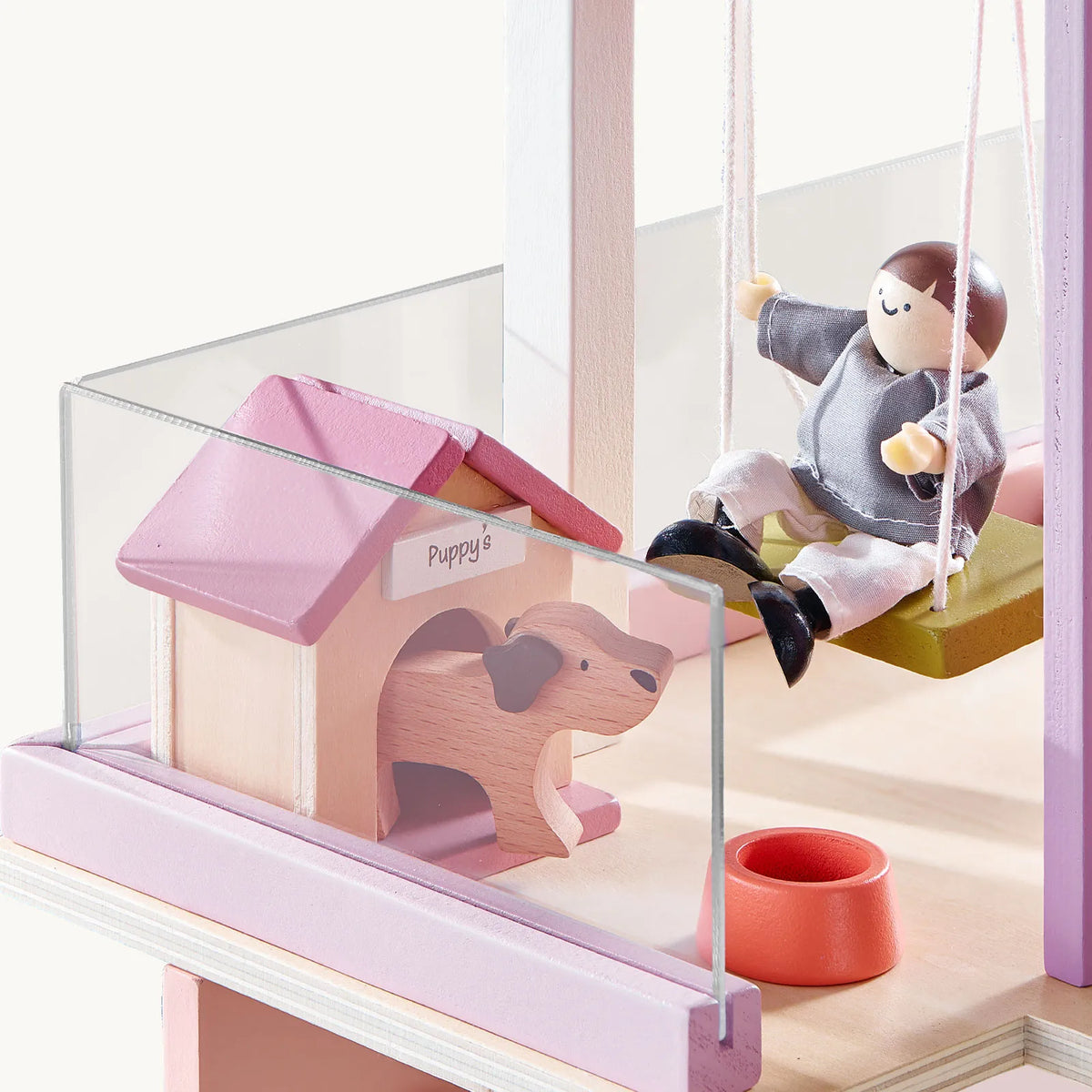 A wooden doll in a gray shirt sits on a 1:12 scale Sweetwood Cottage Dollhouse (48-pc Furniture) swing next to a small dog house labeled Puppys containing a wooden dog figure.