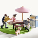 Wooden dollhouse family figures and a dog gather at a 1:12 scale Sweetwood Cottage Dollhouse (48-pc Furniture) picnic table with a pink umbrella and a toy grill.