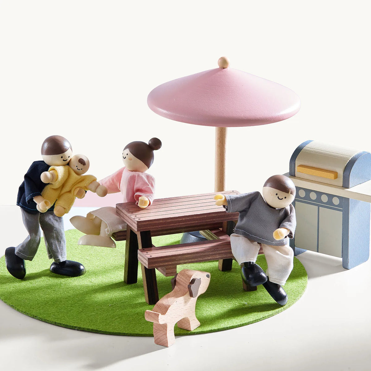 Wooden dollhouse family figures and a dog gather at a 1:12 scale Sweetwood Cottage Dollhouse (48-pc Furniture) picnic table with a pink umbrella and a toy grill.
