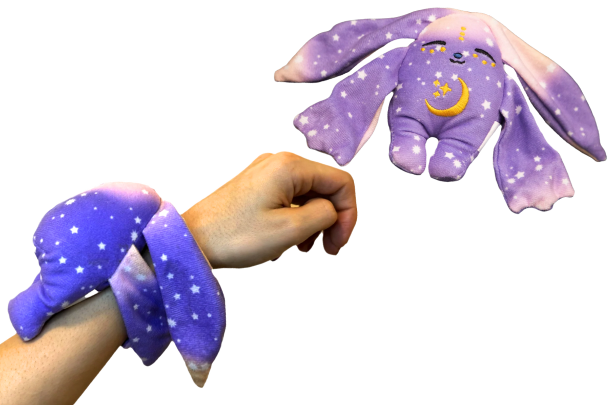 A Polli Mini Wrist Hugger – Magnetic Sensory Plush, a small purple, star-patterned magnetic toy with long ears and a crescent moon chest, shown floating in the air and wrapped around a wrist as a pocket-sized comfort.
