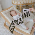 A baby in white lies in a cushioned wooden rocker, gazing at a fold-out high-contrast card from the Tiny Land® Montessori Toys for Newborns (0–3 Months) set. A wicker chair is visible in the background.