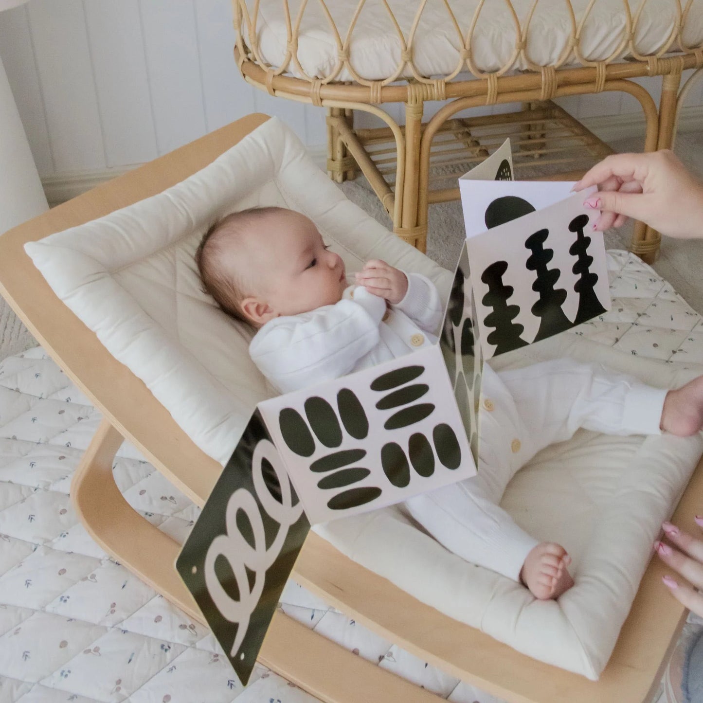 A baby in white lies in a cushioned wooden rocker, gazing at a fold-out high-contrast card from the Tiny Land® Montessori Toys for Newborns (0–3 Months) set. A wicker chair is visible in the background.